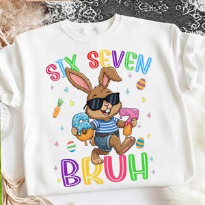Pode incluir: Camisola branca com um coelho de desenho animado usando óculos de sol e segurando um donut em forma do número 6 e um donut em forma do número 7. As palavras "SIX SEVEN BRUH" estão em letras coloridas.