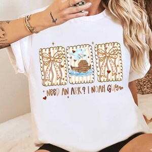 Puede incluir: Camiseta blanca con un dise&ntilde;o gr&aacute;fico de tres marcos rectangulares. Cada marco tiene un borde festoneado y presenta un lazo, un coraz&oacute;n y una representaci&oacute;n del Arca de No&eacute;. El texto "NEED AN ARK? I NOAH GUY" est&aacute; impreso debajo.
