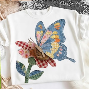 Puede incluir: Sudadera blanca de cuello redondo con un gran aplique de mariposa y flor. La mariposa tiene alas de mezclilla azul con detalles de patchwork en rosa, amarillo y naranja. La flor tiene pétalos de cuadros rojos y rosas y hojas verdes.