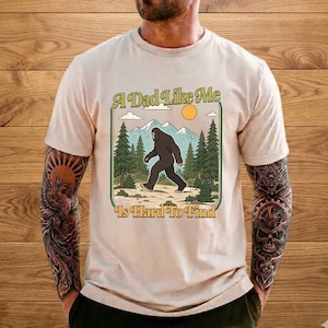 Può includere: T-shirt beige con grafica Bigfoot che cammina in una foresta, con la scritta "A Dad Like Me Is Hard To Find". Il design include montagne, alberi, un sole e nuvole.