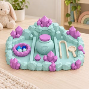 Peut inclure: Un ensemble de jeu sensoriel en plastique bleu clair et violet représentant un paysage rocheux avec des formations de cristaux moulées. Il comprend un pot central avec couvercle orné d'un diamant, un bol avec des gemmes, une pince et une loupe.