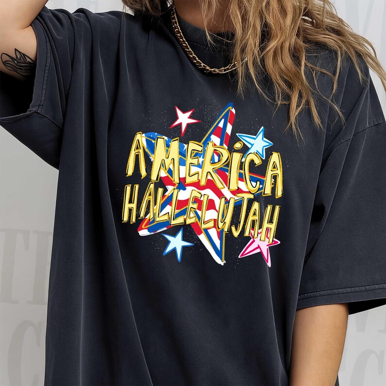 May include: Dark gray t-shirt featuring a star-shaped graphic with the text "AMERICA HALLELUJAH" in yellow. The star design incorporates red, white, and blue elements, with additional star accents in various colors.