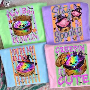 May include: Four t-shirts in light green, purple, light blue, and pink, each featuring a graphic of a colorful dumpling in a bamboo steamer. The designs include Halloween-themed text like Hey Boo Dumplin, Stay Spooky, You're My Boo, and Creepin' It's Cute.