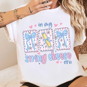 Könnte beinhalten: Weißes T-Shirt mit einem verspielten Design. Das Shirt zeigt den Text "in my string cheese era" in Rosa und Blau, zusammen mit Illustrationen von Schleifen, Blumen und Käse-Sticks.