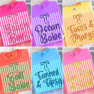 Op de afbeelding: Zes crewneck T-shirts in verschillende kleuren, elk met een uniek ontwerp. De ontwerpen bevatten zinnen als "Beach Bum", "Ocean Babe" en "Tacos & Margs", met gestreepte achtergronden en strikaccenten.