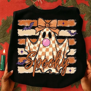 May include: A black sweatshirt featuring a graphic of a ghost with a checkered pattern, wearing an orange bow and blowing a pink bubble. The design includes horizontal orange stripes, purple bats, small stars, and the word Spooky in orange script.