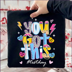 May include: Black t-shirt featuring the phrase "YOU GOT THIS" in a vibrant, playful design. The text incorporates pencil and ruler motifs, lightning bolts, and heart-shaped details. The hashtag "#testday" is visible at the bottom.