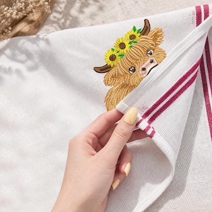 May include: White kitchen towel with an embroidered Highland cow wearing a sunflower crown. The towel has red stripes along one edge. The cow has brown fur and a pink nose. The towel is being held by a person with yellow nail polish.