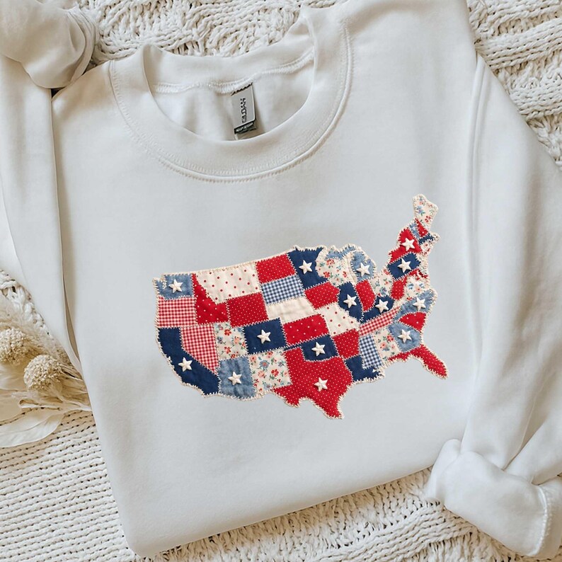 May include: White sweatshirt featuring a patchwork design of the United States map. The map is composed of red, white, and blue fabric squares with star and floral patterns. The sweatshirt has a crew neck and long sleeves.
