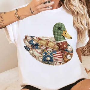 Op de afbeelding: Wit T-shirt met een patchwork eendenontwerp. Het lichaam van de eend bestaat uit honkbalthema patches, waaronder honkballen, knuppels en een Amerikaanse vlag. De kop van de eend is groen en de snavel is geel.
