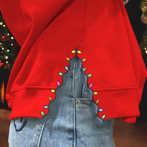 May include: A red sweatshirt with a Christmas tree design made of colorful lights. The tree is outlined on the sweatshirt with a blue denim fabric.