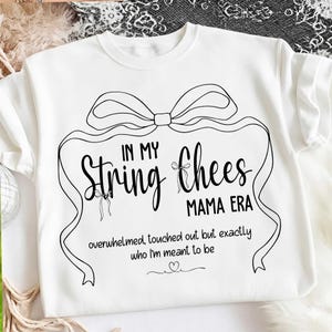 Peut inclure: Sweat-shirt blanc à col rond avec un motif noir. Le motif comprend le texte "IN MY String Chees MAMA ERA" avec un motif de nœud et de ruban. En dessous, la phrase "overwhelmed, touched out but exactly who I'm meant to be."