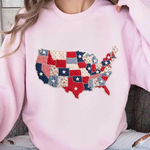 May include: A light pink sweatshirt featuring a patchwork design of the United States map. The map is composed of red, white, and blue fabric squares, some with star accents. The sweatshirt has long sleeves and a crew neck.