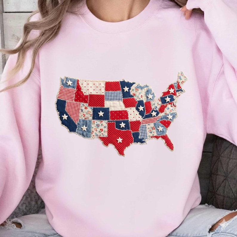 May include: A light pink sweatshirt featuring a patchwork design of the United States map. The map is composed of red, white, and blue fabric squares, some with star accents. The sweatshirt has long sleeves and a crew neck.