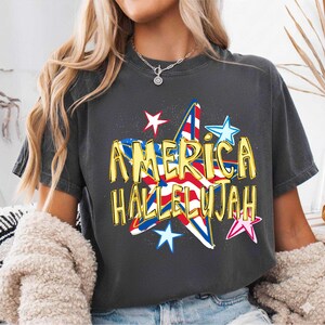 May include: Dark gray t-shirt with the words "AMERICA HALLELUJAH" in gold lettering. The design features a star with red, white, and blue stripes and stars, evoking patriotic themes.
