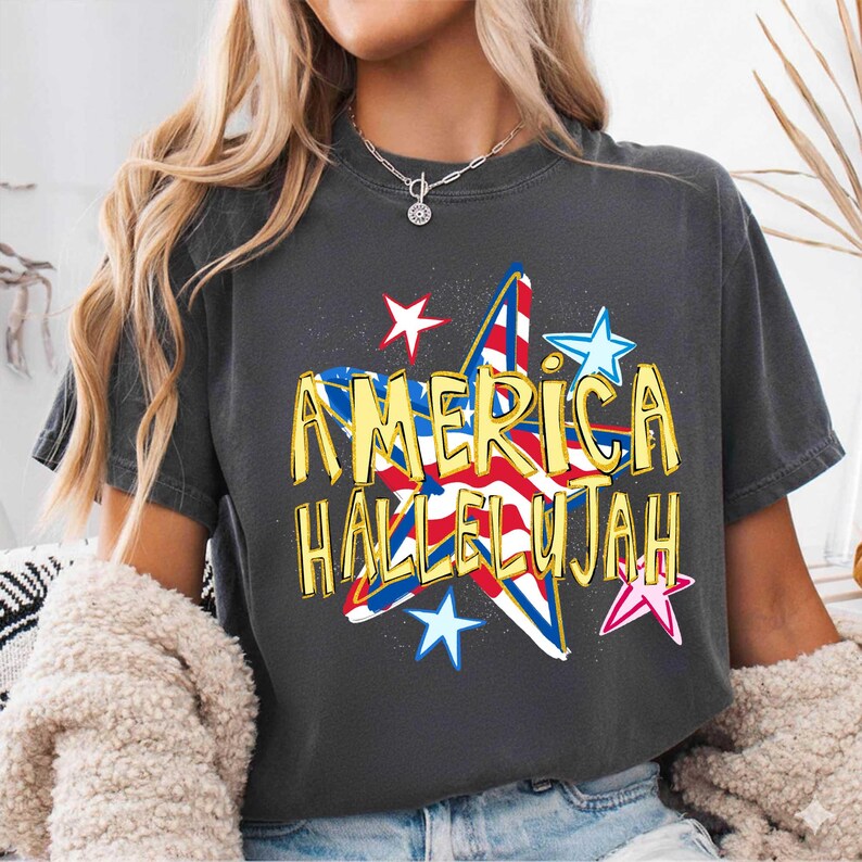 May include: Dark gray t-shirt with the words "AMERICA HALLELUJAH" in gold lettering. The design features a star with red, white, and blue stripes and stars, evoking patriotic themes.