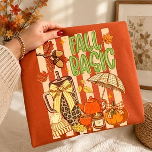 May include: An orange sweatshirt with the words "FALL BASIC" in green and white. The design includes fall-themed illustrations such as a coffee cup, teapot, pumpkin, and umbrella. The sweatshirt is held by a person.