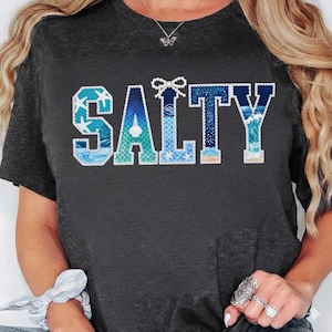 May include: Dark gray t-shirt with the word "SALTY" in a decorative font. The letters are filled with ocean-themed patterns, including starfish, waves, and mermaid scales. A bow detail sits above the "T".