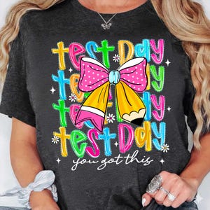 May include: Dark gray t-shirt with a colorful graphic that says "Test Day" multiple times, with a pencil and bow design. The text "you got this" is written in cursive below the graphic. The design includes pink, yellow, blue, and green colors.