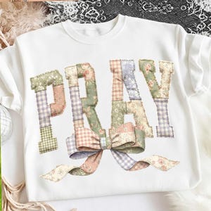 May include: White sweatshirt featuring the word "PRAY" in patchwork lettering. The letters are crafted from floral and gingham fabrics in pastel shades. A large bow, made from matching fabric, adorns the design.