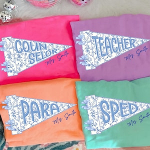 May include: Four brightly colored t-shirts: pink, purple, orange, and green. Each shirt features a pennant design with a floral pattern and a profession title in blue. The titles read: COUNSELOR, TEACHER, PARA, and SPED. The name "Mrs. Smith" is printed below each title.
