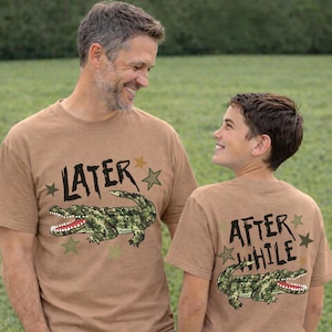 May include: Two tan t-shirts featuring alligator designs. One shirt displays "LATER" with an alligator graphic, the other says "AFTER WHILE" with a matching alligator. Both shirts have star accents.