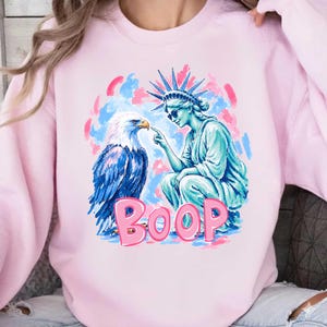 May include: A pale pink sweatshirt with a graphic print. The design features the Statue of Liberty and an eagle, with the word "BOOP" in pink bubble letters. The background has blue and pink brushstrokes.