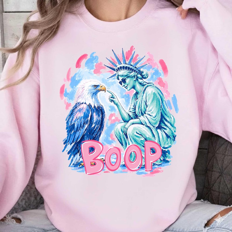 May include: A pale pink sweatshirt with a graphic print. The design features the Statue of Liberty and an eagle, with the word "BOOP" in pink bubble letters. The background has blue and pink brushstrokes.