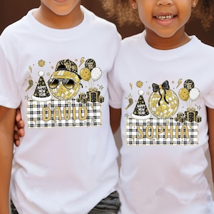 May include: White t-shirts with a New Year's theme. One shirt features a disco ball wearing sunglasses and a hat, with the name "DAVID" on a plaid banner. The other shirt has a disco ball with a bow and the name "SOPHIA".