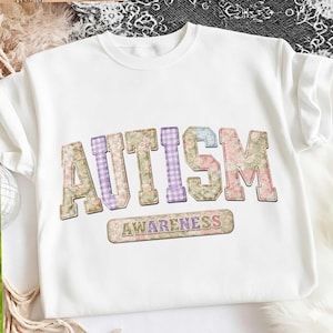 以下が含まれることがあります： 大きな花柄の文字で「AUTISM」と書かれた白いスウェットシャツ。その下に「AWARENESS」と書かれた小さなバナーがあります。スウェットシャツはクルーネックです。