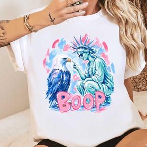 May include: White t-shirt with a graphic of the Statue of Liberty and an eagle. The Statue of Liberty is pointing at the eagle, with the word "BOOP" in pink letters below. The background has pink and blue brushstrokes.