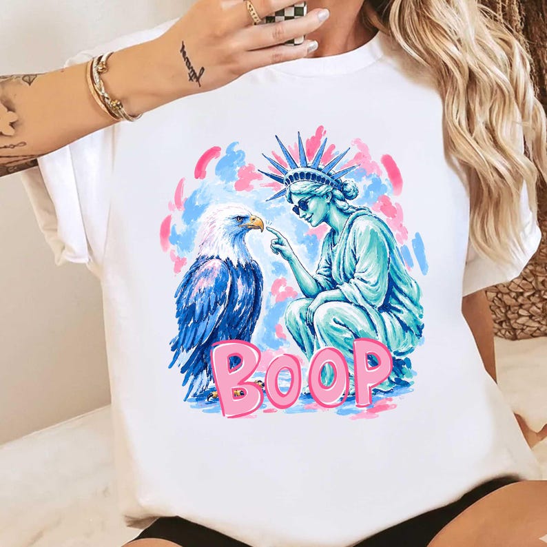 May include: White t-shirt with a graphic of the Statue of Liberty and an eagle. The Statue of Liberty is pointing at the eagle, with the word "BOOP" in pink letters below. The background has pink and blue brushstrokes.