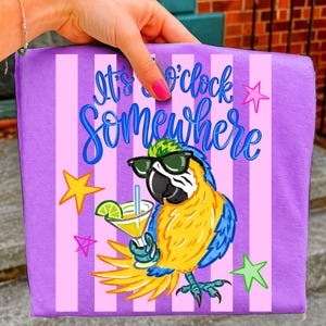 May include: A purple tote bag with pink vertical stripes and the phrase "It's o'clock somewhere" in blue script. A cartoon parrot wearing sunglasses and holding a cocktail is featured on the front, with star accents.