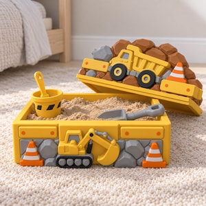 May include: A yellow construction-themed sandbox container with a matching lid. The lid features a moulded dump truck and rocks. The base displays a moulded excavator and traffic cones. Inside, the sandbox contains sand, a yellow bucket, and a grey shovel.