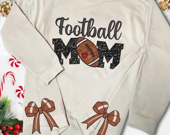 May include: Cream-coloured sweatshirt with the words "Football MOM" in black and glittery black letters. A football with a heart is in the centre. Brown football-shaped bows are on the sleeves. Festive holiday decorations are in the background.