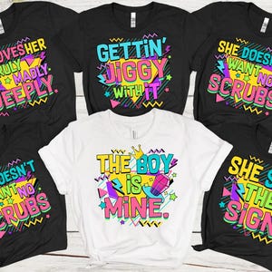 Könnte beinhalten: Satz von sechs schwarzen und weißen T-Shirts mit bunten Grafiken und Texten im Stil der 90er Jahre. Die Shirts zeigen Sätze wie "He Loves Her Truly Madly Deeply", "Gettin' Jiggy With It", "She Doesn't Want No Scrubs", "The Boy Is Mine" und "She Saw The Sign".