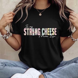Könnte beinhalten: Schwarzes T-Shirt mit dem Text "In my STRING CHEESE Mama Era" in einem farbenfrohen, floralen Design. Das Shirt hat einen Rundhalsausschnitt.