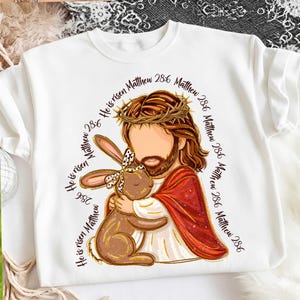 May include: White sweatshirt featuring a graphic of Jesus embracing a brown bunny wearing a bow. Jesus is depicted with a crown of thorns and a red robe. Text around the image reads "He is risen Matthew 28:6".