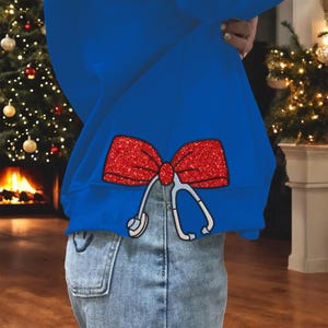 May include: A royal blue sweatshirt with a red glitter bow and a stethoscope design. The sweatshirt is paired with light blue denim jeans. The image is set against a backdrop of a Christmas tree and a fireplace.