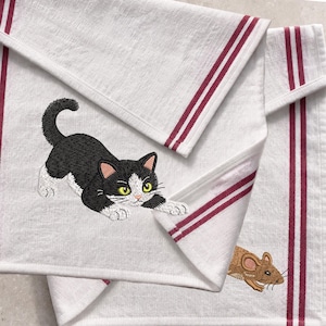 May include: White kitchen towels featuring embroidered cat and mouse motifs. The cat is black and white with yellow eyes, and the mouse is brown. The towels have red stripes along the edges, providing a contrasting colour to the design.