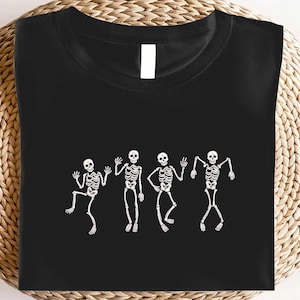 May include: Black t-shirt featuring a white graphic of four dancing skeletons. The skeletons are in various poses, with arms raised and legs bent, giving a playful and festive vibe. Perfect for Halloween or any occasion.