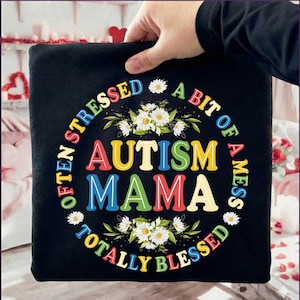 Puede incluir: Camiseta negra con el texto "Often Stressed, A Bit of a Mess, Autism Mama, Totally Blessed" en letras coloridas. El diseño incluye margaritas blancas y hojas verdes, creando un acento floral.