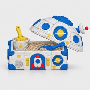 May include: A white and blue space-themed sandbox container with a hinged dome lid. The base features a red and white rocket ship, yellow stars, and planet motifs. Inside, a small white bucket with a yellow rake and a blue shovel rest in sand.