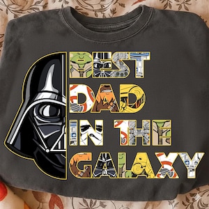 May include: Dark gray sweatshirt featuring Darth Vader's helmet on the left and the text "BEST DAD IN THE GALAXY" in a colorful, Star Wars-themed font. The letters are filled with various character designs and patterns.