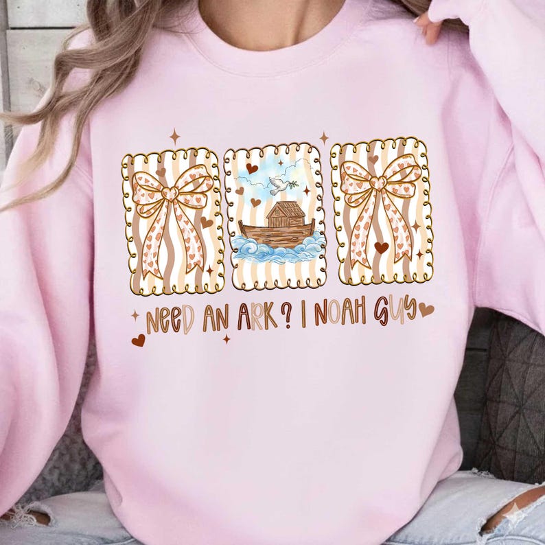 Puede incluir: Sudadera rosa claro con un gr&aacute;fico que presenta tres im&aacute;genes enmarcadas. Las im&aacute;genes incluyen un lazo, una representaci&oacute;n del Arca de No&eacute; y otro lazo. El texto "NEED AN ARK? I NOAH GUY" est&aacute; impreso debajo.