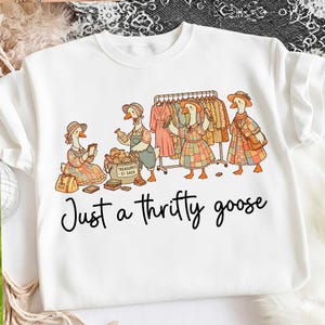 Puede incluir: Sudadera blanca con una ilustración caprichosa de gansos comprando en una tienda de segunda mano. Los gansos llevan sombreros y vestidos, y miran la ropa en una percha y una caja de tesoros. El texto "Just a thrifty goose" está debajo.
