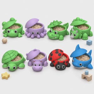 Può includere: Una collezione di otto contenitori a forma di animale con coperchio, ciascuno riempito con una sostanza granulare di colore chiaro. Il set include una rana verde, un polpo viola, un coccodrillo verde, un triceratopo verde, una tartaruga verde, un ippopotamo viola, una coccinella rossa e uno squalo blu.