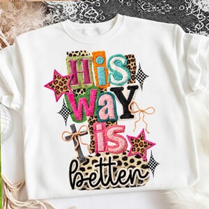 May include: White sweatshirt with the phrase "His Way is Better" in colorful, textured letters. The design includes stars and bows, with leopard print accents. A casual, graphic tee.