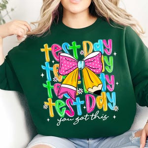 May include: Dark green sweatshirt with a colorful graphic. The graphic includes the words "Test Day" in multiple colors, a pink polka dot bow, and a yellow pencil. The phrase "you got this" is also included.