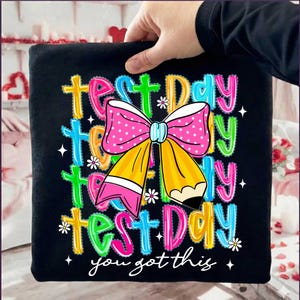 May include: Black t-shirt with a colorful graphic that says "Test Day" multiple times, along with a pink polka-dot bow and a yellow pencil. The text "you got this" is at the bottom, with small white flowers and stars.